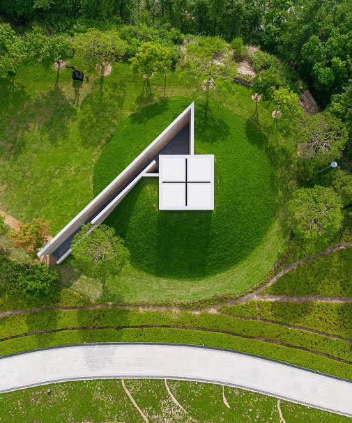 Aerial view of a concrete Tadao Ando chapel with cruciform roof opening set into a green hillside